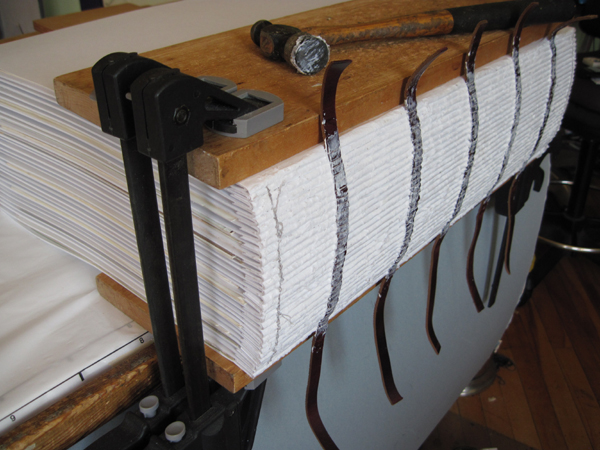 large handbound book in clamp after hand sewing, gluing and hammering the spine into a rounded shape. sewing around leather cords.